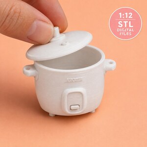 Puede incluir: Una arrocera miniatura de color blanco con tapa extraíble, que presenta la palabra Aroma grabada en el frente y un pequeño interruptor. El objeto es un archivo digital a escala 1:12 para impresión 3D, presentado como un modelo de plástico.
