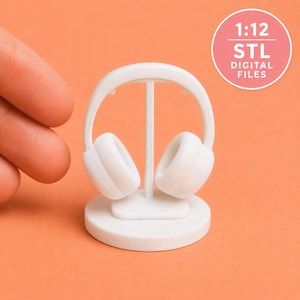 May include: A white 3D printed miniature headphone set on a circular stand. The text 1:12 STL DIGITAL FILES is printed in white on a pink circular background. The headphones feature a curved headband and two ear cups attached to a central vertical support.
