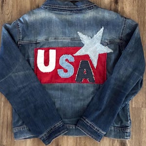 May include: A denim jacket with a patriotic design. The back features a red rectangle with the letters "USA" in white, light blue, and navy blue. A light blue star is above the letters. The jacket is made of blue denim.