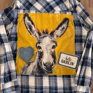 May include: A blue and white plaid long-sleeve button-down shirt featuring a large mustard yellow square patch on the back. The patch displays a painted donkey, a denim heart, and a rectangular fabric label with the text hello DARLIN'.