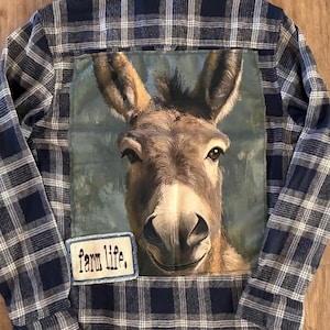 May include: A long-sleeve button-down shirt featuring a navy blue and white plaid pattern. The back displays a large, rectangular graphic of a donkey's face and a smaller rectangular patch with the text "farm life." in black lettering.