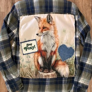 May include: A long-sleeve button-down shirt in a blue, green, and white plaid pattern. The back features a large rectangular fabric patch with a detailed illustration of a fox sitting on a stump, a blue denim heart patch, and a small rectangular patch with the text Stay Foxy!