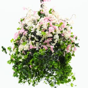 Könnte beinhalten: Ein hängendes Blumenarrangement mit rosa und weißen Blüten und grünem Laub. Das Arrangement hängt an einem dunklen, strukturierten Stiel vor einem weißen Hintergrund. Die Blüten und das Laub sind dicht gepackt.