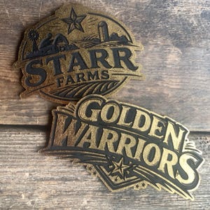 May include: Two die-cut stickers with a gold glitter finish and black lettering. The top sticker features a farm scene with a barn, silo, and windmill, labeled STARR FARMS. The bottom sticker displays the text GOLDEN WARRIORS with a star.