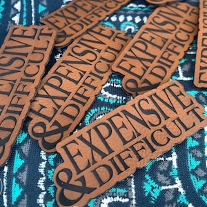 May include: Several brown rectangular patches with the words "EXPENSIVE & DIFFICULT" in black lettering. The patches are scattered on a patterned blue and white fabric. The patches have a rounded bottom edge.
