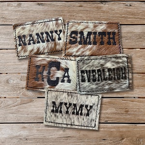 May include: Five rectangular patches with stitched edges and various text. The patches are made of cowhide with a natural pattern of brown, white, and black. The text includes "NANNY", "SMITH", "HCA", "EVERLEIGH", and "MYMY" in black.