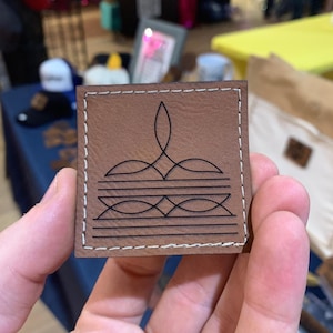 May include: A square brown leather patch featuring white perimeter stitching and a black laser-engraved design consisting of a central pointed flourish above two horizontal lines and a geometric pattern of intersecting lines and diamond shapes.