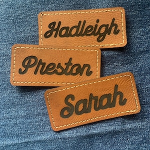 May include: Three brown leather name tags with black cursive lettering. The names Hadleigh, Preston, and Sarah are displayed. Each tag has a stitched border and is set against a blue denim background.