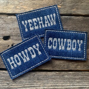 K&ouml;nnte beinhalten: Drei Denim-Patches mit wei&szlig; gesticktem Text. Die Patches lesen "YEEHAW", "COWBOY" und "HOWDY". Jeder Patch hat einen wei&szlig; gen&auml;hten Rand. Die Patches sind auf einer rustikalen Holzoberfl&auml;che angeordnet.