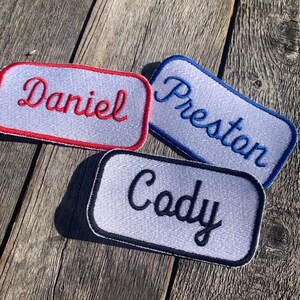 May include: Three embroidered name patches: "Daniel" in red, "Preston" in blue, and "Cody" in black. Each name is stitched in a cursive font on a white rectangular patch with a colored border. The patches are displayed on a weathered wooden surface.