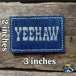 K&ouml;nnte beinhalten: Ein rechteckiger Denim-Aufn&auml;her mit dem Wort "YEEHAW" in Wei&szlig; gestickt. Der Aufn&auml;her ist ca. 7,6 cm lang und 5,1 cm breit, mit einem wei&szlig;en, gen&auml;hten Rand. Er liegt auf einem rustikalen Holzgrund.