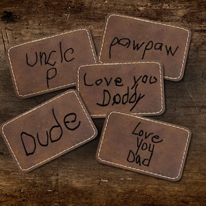 May include: Five brown leather patches with white stitching and handwritten text. The patches say "Uncle P", "Dude", "Pawpaw", "Love you Doddy", and "Love You Dad". The patches are arranged on a wooden surface.