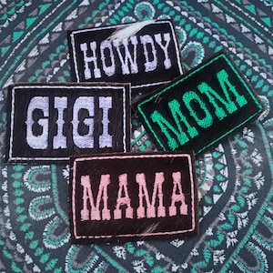 May include: Four rectangular patches with embroidered text. The patches are black, brown, and green, with white and pink lettering. The text includes "HOWDY", "GIGI", "MOM", and "MAMA". A feather is visible on one patch.