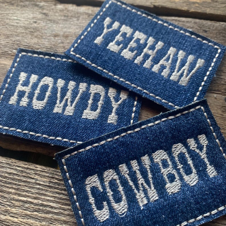 K&ouml;nnte beinhalten: Drei blaue Denim-Patches mit wei&szlig; gesticktem Text. Die Patches lesen "HOWDY", "YEEHAW" und "COWBOY". Jeder Patch hat einen wei&szlig; gen&auml;hten Rand.