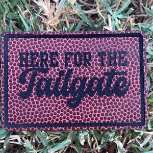 May include: A rectangular patch with a football texture background and the words "HERE FOR THE Tailgate" in black lettering. The patch has a black border and is set against a backdrop of green grass.