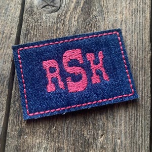K&ouml;nnte beinhalten: Ein marineblauer, gestickter Aufn&auml;her mit den Buchstaben "RSH" in leuchtendem Pink. Der Aufn&auml;her ist rechteckig mit rosa N&auml;hten an den R&auml;ndern. Der Aufn&auml;her liegt auf einer Holzoberfl&auml;che.
