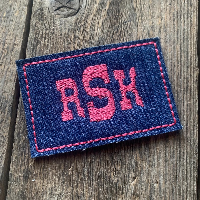 K&ouml;nnte beinhalten: Ein marineblauer, gestickter Aufn&auml;her mit den Buchstaben "RSH" in leuchtendem Pink. Der Aufn&auml;her ist rechteckig mit rosa N&auml;hten an den R&auml;ndern. Der Aufn&auml;her liegt auf einer Holzoberfl&auml;che.