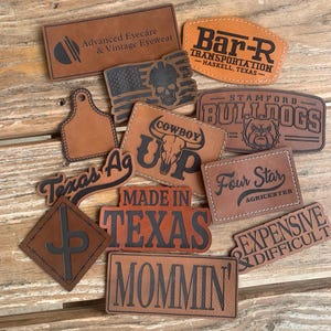 May include: A collection of tan and brown leather patches featuring various laser-engraved designs and text including Advanced Eyecare, Bar-R Transportation, Stamford Bulldogs, Cowboy Up, Texas Ag, Made in Texas, Four Star Agricenter, and Mommin'.