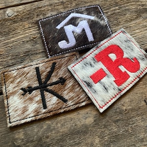 May include: Three rectangular patches made of textured animal hide with hair. One dark brown patch features a white embroidered JM logo. A tan and white patch displays a black embroidered arrow symbol. A white and black patch shows a red embroidered -R.