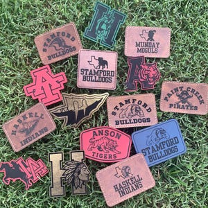 May include: A collection of leather patches featuring various school mascots and team names. The patches are in various colors, including brown, red, blue, and green. The patches include the names "Stamford Bulldogs", "Haskell Indians", and "Anson Tigers".