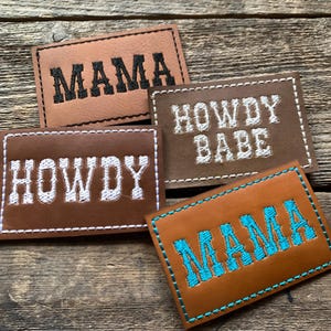 May include: Four brown leather patches with embroidered text. Two patches read "MAMA", one in black and one in turquoise. The other two patches read "HOWDY" and "HOWDY BABE" in white. All patches have white or turquoise stitching.