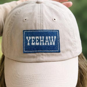 K&ouml;nnte beinhalten: Beige Baseball Cap mit gebogenem Schirm. Die Kappe hat einen blauen Denim-Patch mit dem Wort "YEEHAW" in Wei&szlig; gestickt. Die Kappe ist aus einem weichen Stoff gefertigt und hat einen klassischen, l&auml;ssigen Stil.
