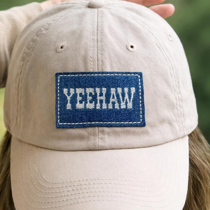 K&ouml;nnte beinhalten: Beige Baseball Cap mit gebogenem Schirm. Die Kappe hat einen blauen Denim-Patch mit dem Wort "YEEHAW" in Wei&szlig; gestickt. Die Kappe ist aus einem weichen Stoff gefertigt und hat einen klassischen, l&auml;ssigen Stil.