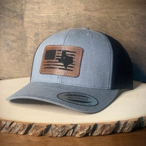 May include: A gray and black trucker hat with a brown leather patch featuring a distressed American flag with the state of Texas superimposed on it. The patch has stitching around the edges. The hat has a black mesh back and a white label that reads "The Authentic Snapback Yupoong Since 1974".