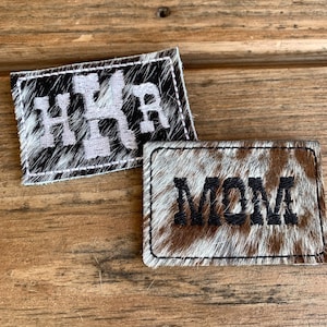 May include: Two rectangular patches with a cowhide pattern. One patch has the letters "HXR" in white, and the other says "MOM" in black. Both patches have a white stitched border.