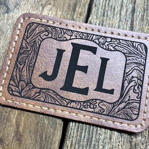 May include: A rectangular, brown leather patch with the initials "JEL" in bold, black letters. The patch features an ornate, floral border and is stitched with a light-colored thread.