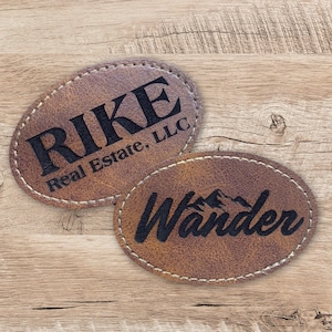 May include: Two brown leather patches with stitched edges. One patch has the text "RIKE Real Estate, LLC" in black lettering. The other patch has the text "Wander" in black lettering with a mountain range graphic.