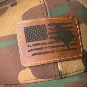 May include: A camouflage trucker hat with a brown leather patch featuring a distressed American flag with the state of Texas superimposed over it.