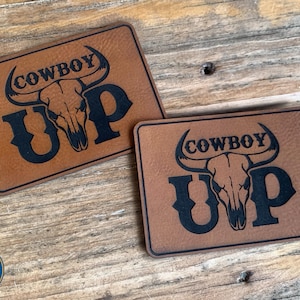 May include: Two rectangular brown leather patches with the words "COWBOY UP" and a bull skull graphic in black. The patches are on a wooden surface. The logo of "TWO SEVENTY SEVEN CREATIVE" is in the bottom left corner.