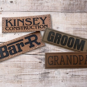 May include: Four rectangular leather patches with embossed text. The patches are in shades of brown and tan, with black lettering. The text includes "KINSEY CONSTRUCTION", "Bar-R", "GROOM", and "GRANDPA". The patches are stitched with white thread.
