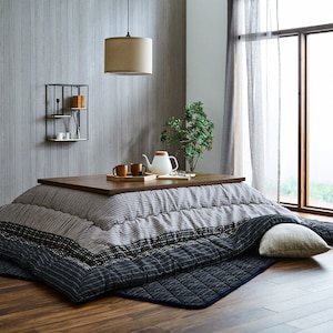 Puede incluir: Un kotatsu japonés tradicional, una mesa baja con una manta calefactada debajo, está instalado en una habitación con suelo de madera. El kotatsu tiene una tapa de madera y un futón a rayas, azul y blanco. Una bandeja con tazas de té y una tetera se encuentra sobre la mesa. Una planta en maceta y una lámpara colgante son visibles en el fondo.