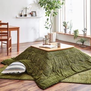 May include: A low, square wooden table with a light wood top sits atop a large, olive green, textured blanket. A striped pillow rests on the blanket. The room has a wooden floor and a large window with plants.
