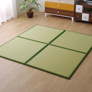 Op de afbeelding: Vier lichtgroene tatami-matten met donkergroene randen zijn gerangschikt op een lichte houten vloer. De matten zijn rechthoekig en vormen een vierkante vorm. Een potplant en meubels staan op de achtergrond.