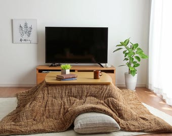 May include: A brown and beige electric heated blanket with a wooden table top and a mug on top of the table. The blanket is on a light beige rug in a room with a television and a plant.