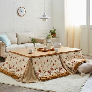 May include: A low, wooden kotatsu table with a patterned blanket featuring red and gold snowflakes. The table is set with cups, a tray, and a teapot. A beige sofa and a white brick wall are in the background.