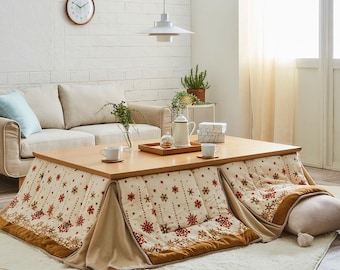 May include: A low, wooden kotatsu table with a patterned blanket featuring red and gold snowflakes. The table is set with cups, a tray, and a teapot. A beige sofa and a white brick wall are in the background.