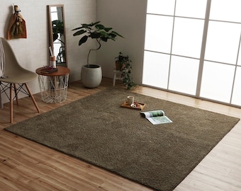 May include: A large, rectangular, brown shag rug dominates the room's wooden floor. The rug is the focal point, with a coffee table, chair, and potted plants arranged around it. A magazine and tray with cups sit on the rug near a large window.