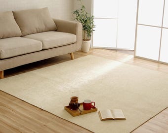 May include: A large, rectangular beige rug in a living room setting. A beige sofa, a small plant, and a window are visible in the background. A wooden tray with a coffee pot, a red mug, and a book are on the rug.