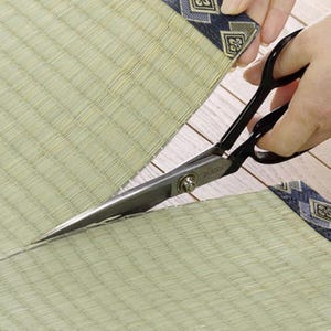 May include: A pair of black scissors with silver blades are cutting a light green woven mat with a blue and white patterned border.