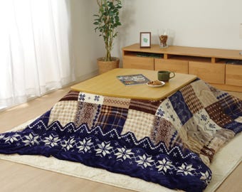 Puede incluir: Un kotatsu eléctrico con calefacción de patrones azules y marrones con una mesa de madera. El kotatsu está sobre una alfombra blanca y hay una planta en una maceta en el fondo.