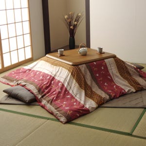 以下が含まれることがあります： 低い木製のこたつテーブルで、上には小さな急須と湯呑みが2つ置かれています。テーブルは赤、茶、クリーム色のストライプ柄の厚手の掛け布団で覆われており、平らな床マットの上に設置されています。