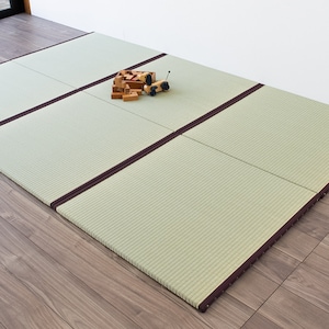 Może przedstawiać: Zestaw czterech jasnozielonych mat tatami z brązowymi obramowaniami. Maty są rozłożone na drewnianej podłodze. Na matach znajduje się mały drewniany samochodzik i stos drewnianych klocków.