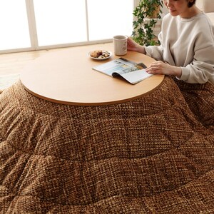 Op de afbeelding: Een ronde futon in bruin en beige met een houten tafelblad in het midden. Op het tafelblad liggen een tijdschrift, een kop koffie en een bord met koekjes.