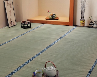 May include: A traditional Japanese room featuring light green tatami floor mats with blue patterned fabric borders. A white ceramic teapot and a matching teacup sit on a dark brown woven tray in the foreground, with a small black stand and floral arrangement visible in the background.