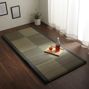 May include: A black and beige woven tatami mat with a black border. The mat is laid out on a wooden floor with a glass of liquid and a bottle on a wooden tray.