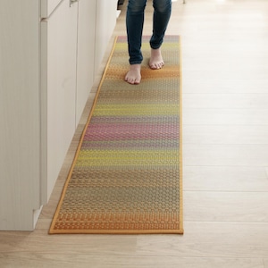 Op de afbeelding: Een lange, rechthoekige loper met een horizontaal streeppatroon in een verloop van warme en koele kleuren, waaronder tinten oranje, geel, roze, groen en bruin. Het kleed heeft een geweven, getextureerd oppervlak en een effen oranje rand.